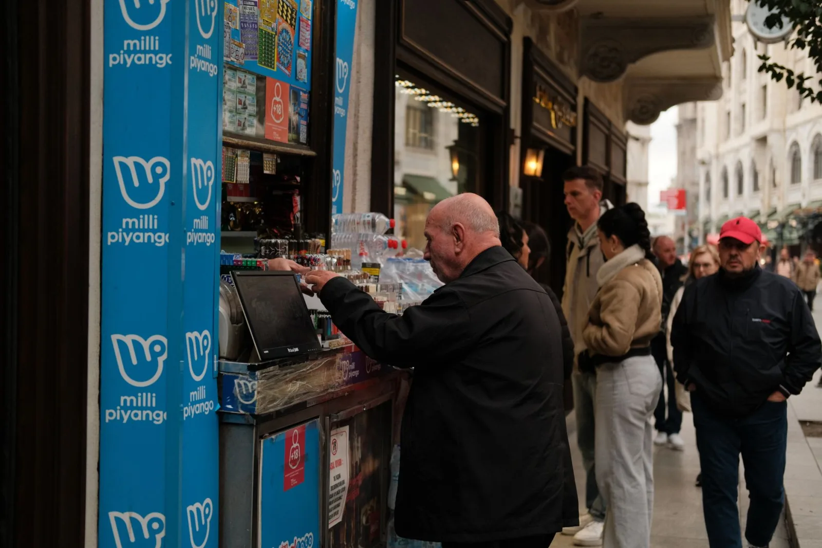 Personas comprando boletos de lotería y bebidas a un vendedor ambulante en una calle urbana concurrida.