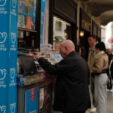 Personas comprando boletos de lotería y bebidas a un vendedor ambulante en una calle urbana concurrida.