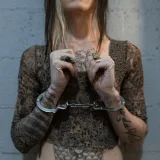 Woman with tattoos and handcuffs, standing against a brick wall, depicting crime and punishment.