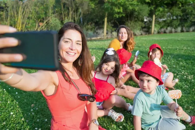 A joyful family picnic with a woman and children taking a group selfie in a sunny park.