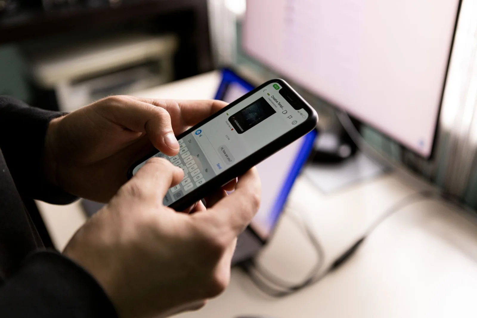 Una persona sosteniendo un smartphone y chateando en interiores cerca de una pantalla de computadora.