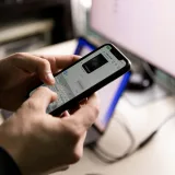 Una persona sosteniendo un smartphone y chateando en interiores cerca de una pantalla de computadora.