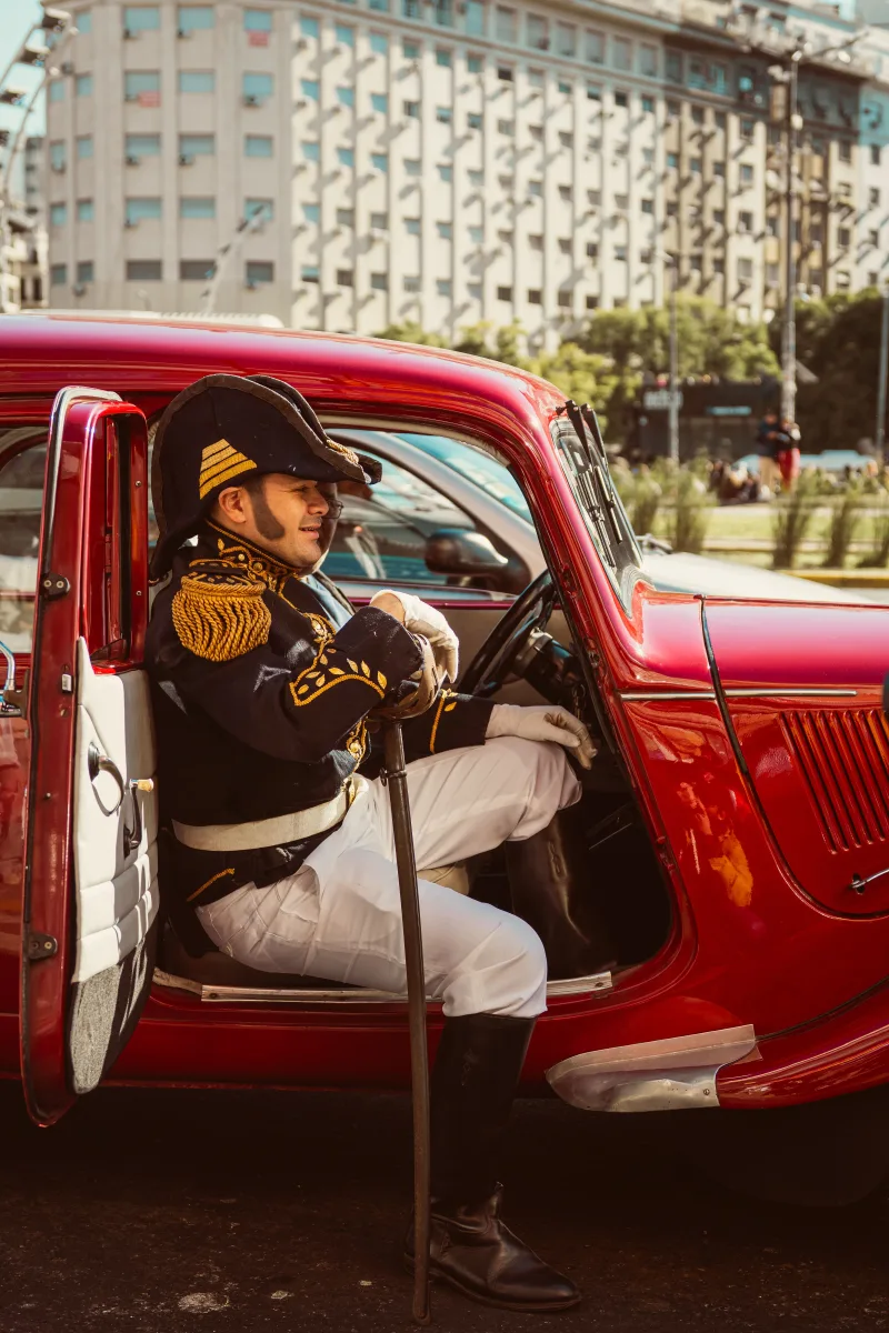 A historic reenactor in uniform sits in a red vintage car on a sunny day in Buenos Aires, Argentina.