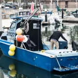Barco de pesca en un animado puerto, con un hombre preparando el equipo, rodeado de embarcaciones.