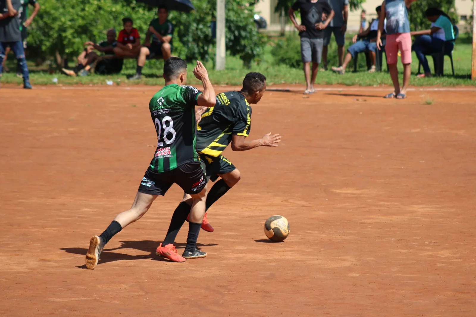 Dos atletas participan en un partido de fútbol competitivo en un campo de tierra ante el público.