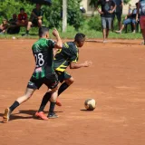 Dos atletas participan en un partido de fútbol competitivo en un campo de tierra ante el público.