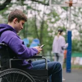 Un joven en silla de ruedas usa un teléfono inteligente en un parque al aire libre.