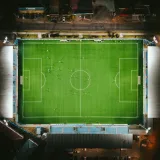 Vista aérea de un estadio de fútbol muy iluminado por la noche, con jugadores en el campo.