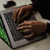 Vista desde arriba de un hacker anónimo escribiendo en una netbook con datos en pantalla mientras está sentado en un escritorio