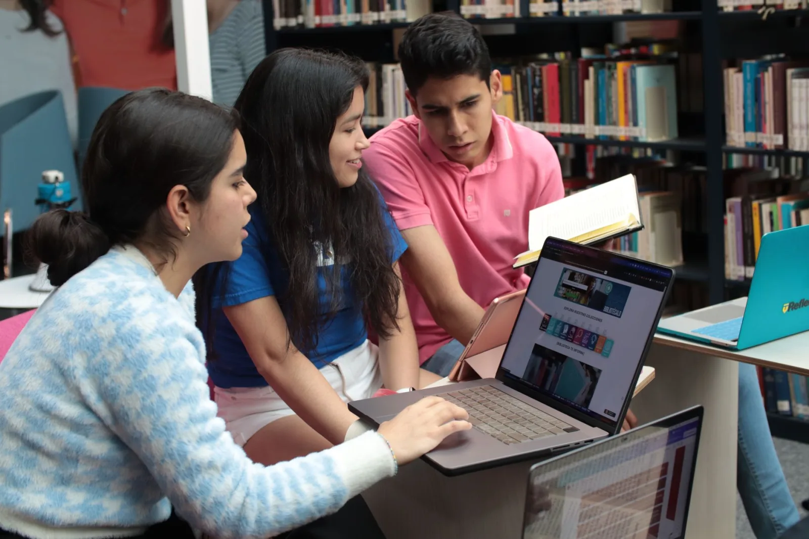 Grupo de jóvenes estudiantes estudiando y conversando en una biblioteca, rodeados de libros y computadoras portátiles.