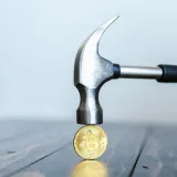 Close-up of a hammer poised above a Bitcoin coin on a wooden surface, representing cryptocurrency challenges.