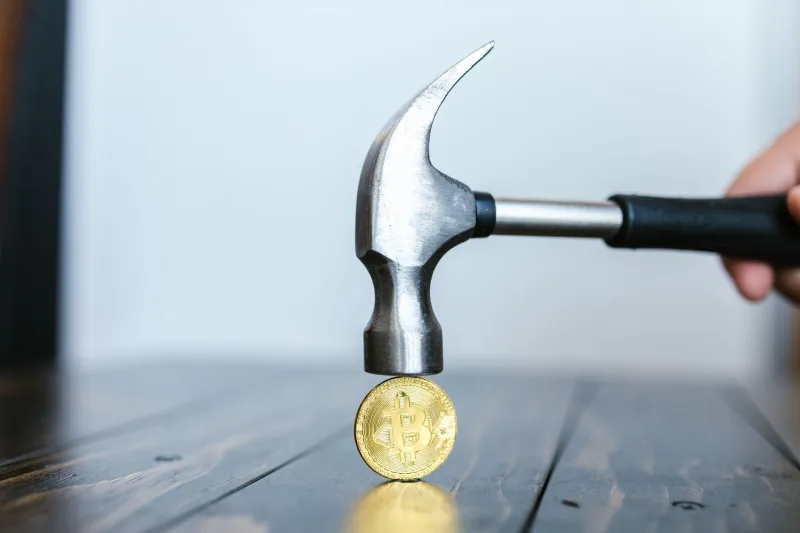 Close-up of a hammer poised above a Bitcoin coin on a wooden surface, representing cryptocurrency challenges.