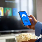 Persona usando una VPN en su smartphone mientras ve la televisión inteligente en casa.