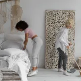 Dos mujeres ordenando un dormitorio contemporáneo con elementos decorativos naturales, mostrando hábitos de limpieza con estilo.