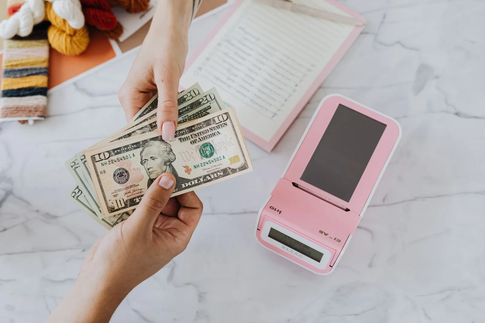 Primer plano de manos intercambiando dólares estadounidenses con una calculadora rosa sobre una superficie de mármol.