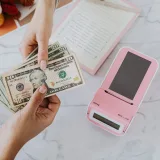 Primer plano de manos intercambiando dólares estadounidenses con una calculadora rosa sobre una superficie de mármol.