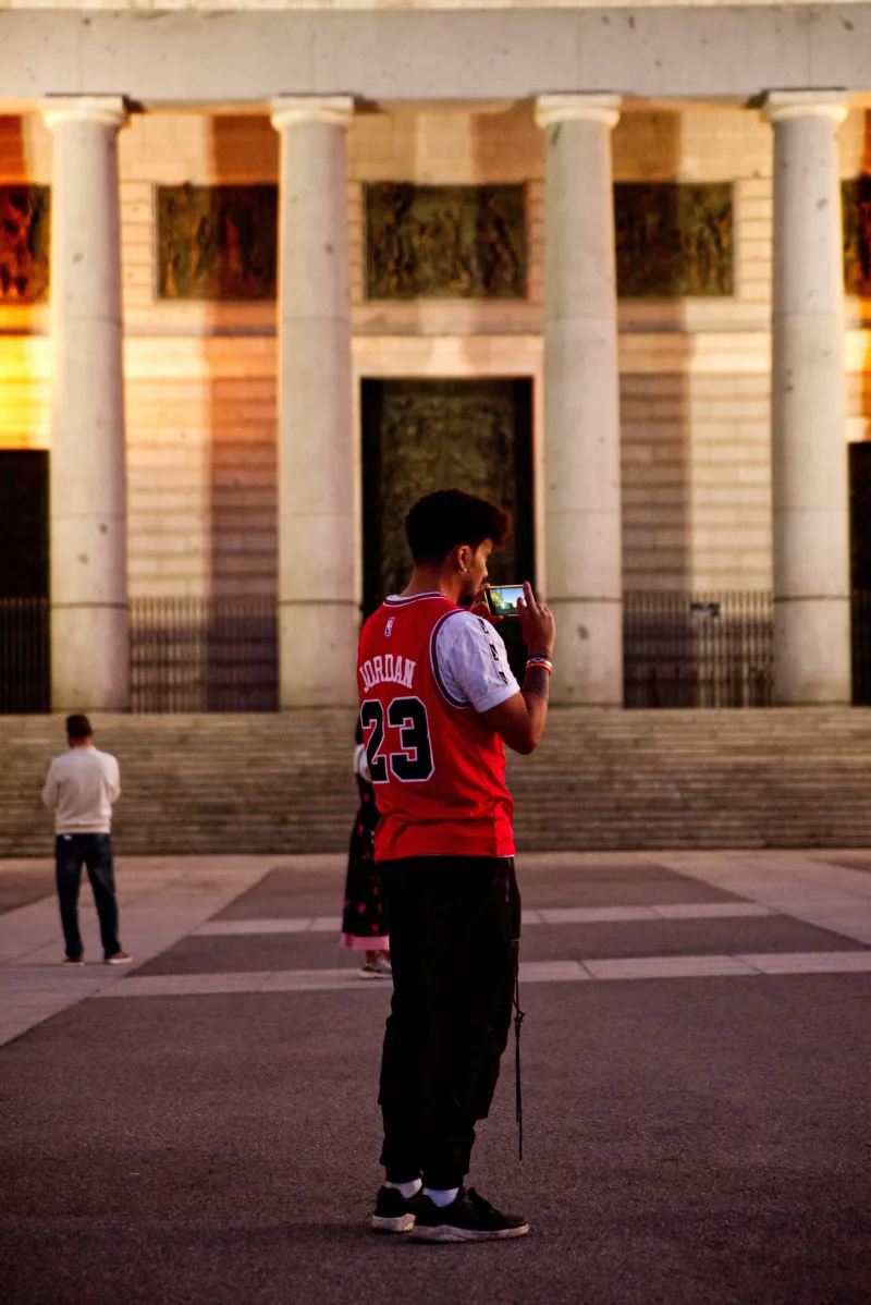 Hombre con camiseta de la selección de Jordania tomando una foto de un edificio histórico en Madrid al atardecer.