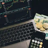 A laptop displaying stock charts with Bitcoin, Euros, and a cellphone calculator, showcasing financial analysis.