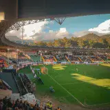 A bustling soccer stadium at sunset with spectators enjoying the game amidst scenic mountains.