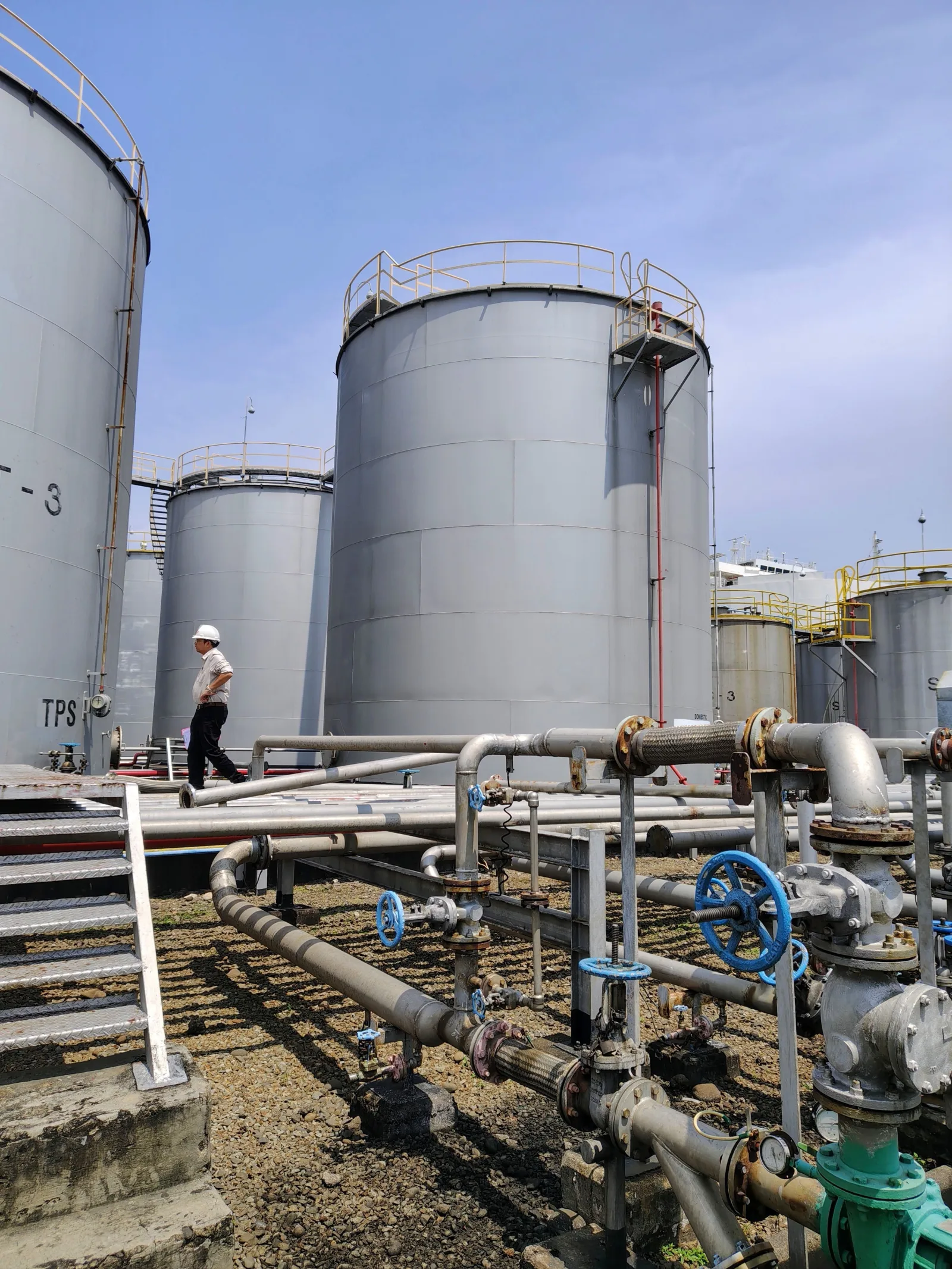 Persona caminando cerca de grandes tanques de almacenamiento de petróleo industriales y tuberías bajo un cielo azul claro.
