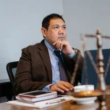 Pensive Asian male lawyer in formal suit browsing netbook while sitting at table with decorative scale of justice and touching chin in contemplation