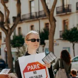 A woman in sunglasses holds a protest sign on a sunny street with trees.