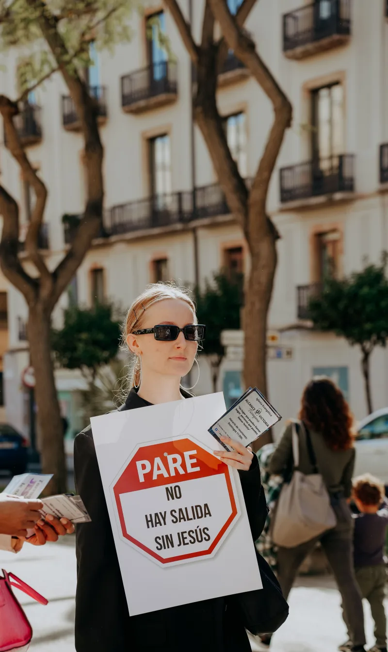 A woman in sunglasses holds a protest sign on a sunny street with trees.