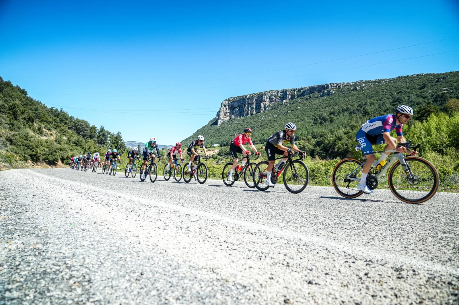 Professional cyclists speed through mountainous terrain in Mersin, Türkiye on a sunny day.