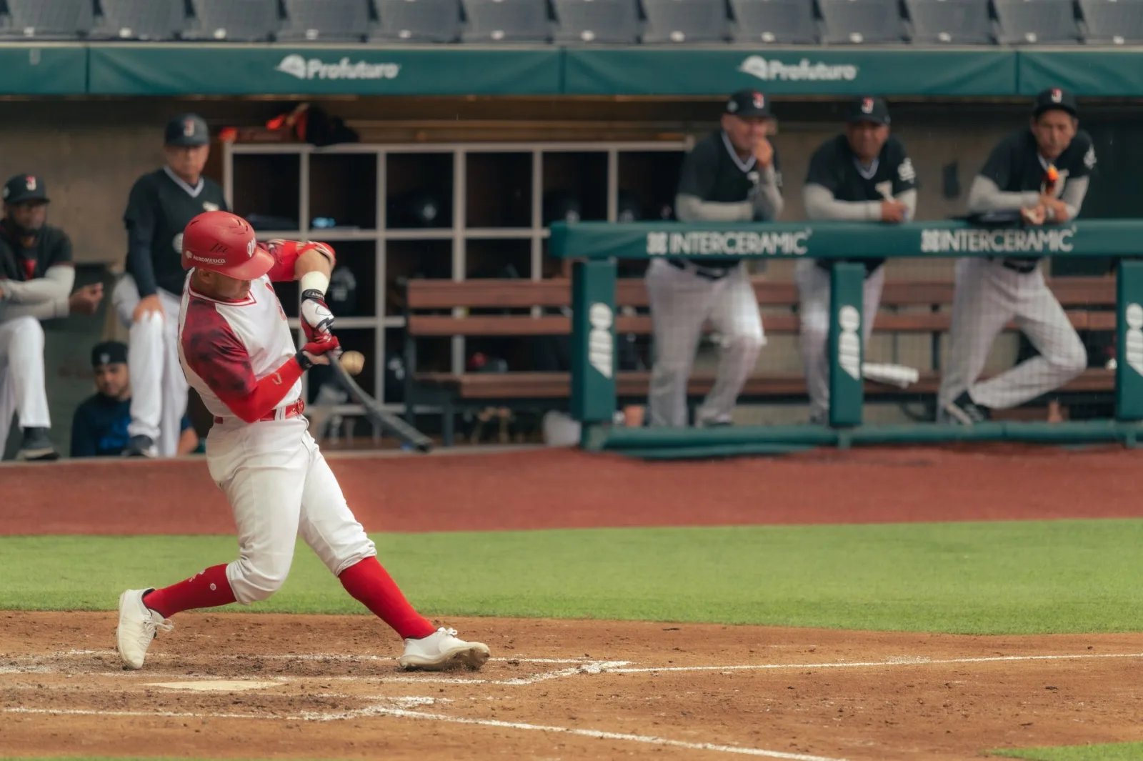 Jugador de béisbol bateando un lanzamiento durante un partido, mientras los entrenadores observan desde el banquillo.