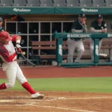 Jugador de béisbol bateando un lanzamiento durante un partido, mientras los entrenadores observan desde el banquillo.