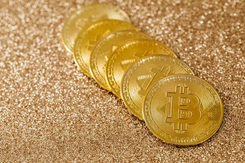 A stack of gold Bitcoin coins placed on a shimmering gold glitter background.