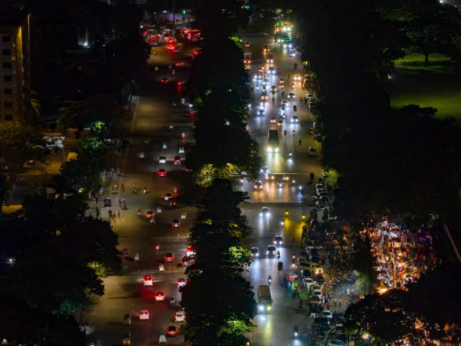 Vista aérea de una animada calle urbana por la noche, con tráfico iluminado y vías bordeadas de árboles.