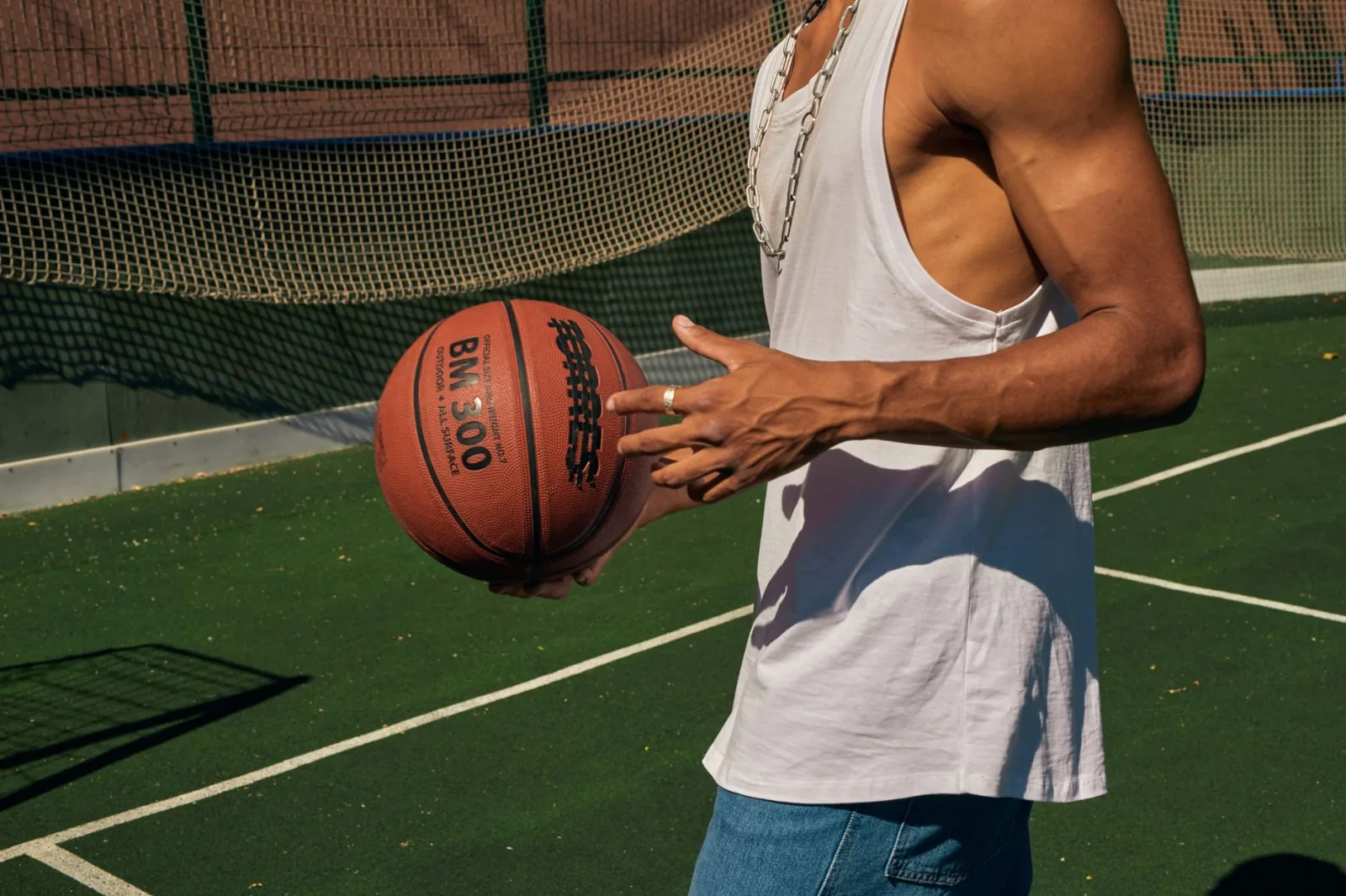 Primer plano de un hombre sosteniendo una pelota de baloncesto en una cancha al aire libre, con una camiseta sin mangas.