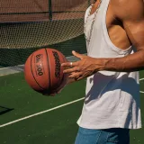 Primer plano de un hombre sosteniendo una pelota de baloncesto en una cancha al aire libre, con una camiseta sin mangas.