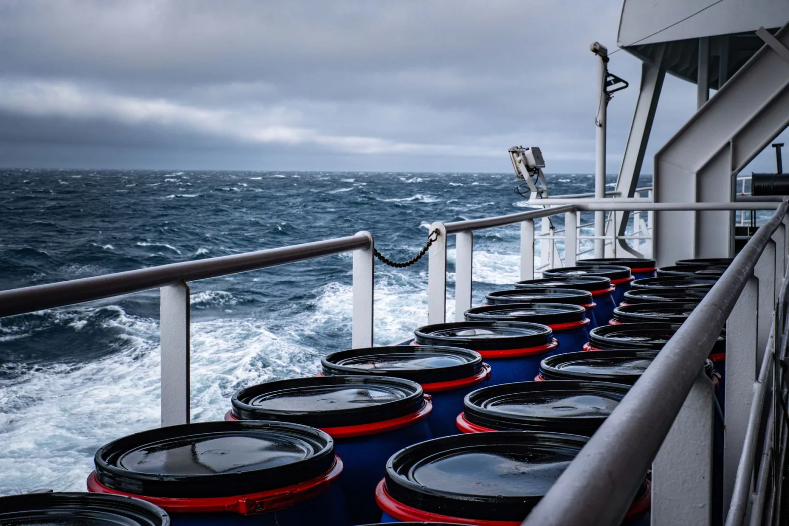 Barriles industriales apilados en la cubierta de un barco en medio de un mar tormentoso, lo que sugiere transporte marítimo.
