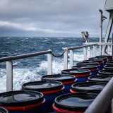 Barriles industriales apilados en la cubierta de un barco en medio de un mar tormentoso, lo que sugiere transporte marítimo.