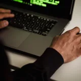 Vista desde arriba de un desarrollador sin rostro, vestido con una sudadera negra con capucha, que escribe código en una netbook mientras trabaja en un estudio luminoso.