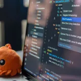 La pantalla de una computadora portátil muestra un editor de código con un adorable peluche de cangrejo naranja a su lado.