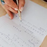 Una persona escribe ecuaciones matemáticas a mano con un lápiz sobre papel cuadriculado, lo que fomenta el aprendizaje.