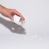 Una mano sostiene un frasco de vidrio y vierte pastillas sobre una superficie blanca en un entorno minimalista.