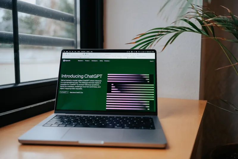 Una laptop que muestra ChatGPT sobre un escritorio junto a una ventana, en un moderno espacio de trabajo en casa.