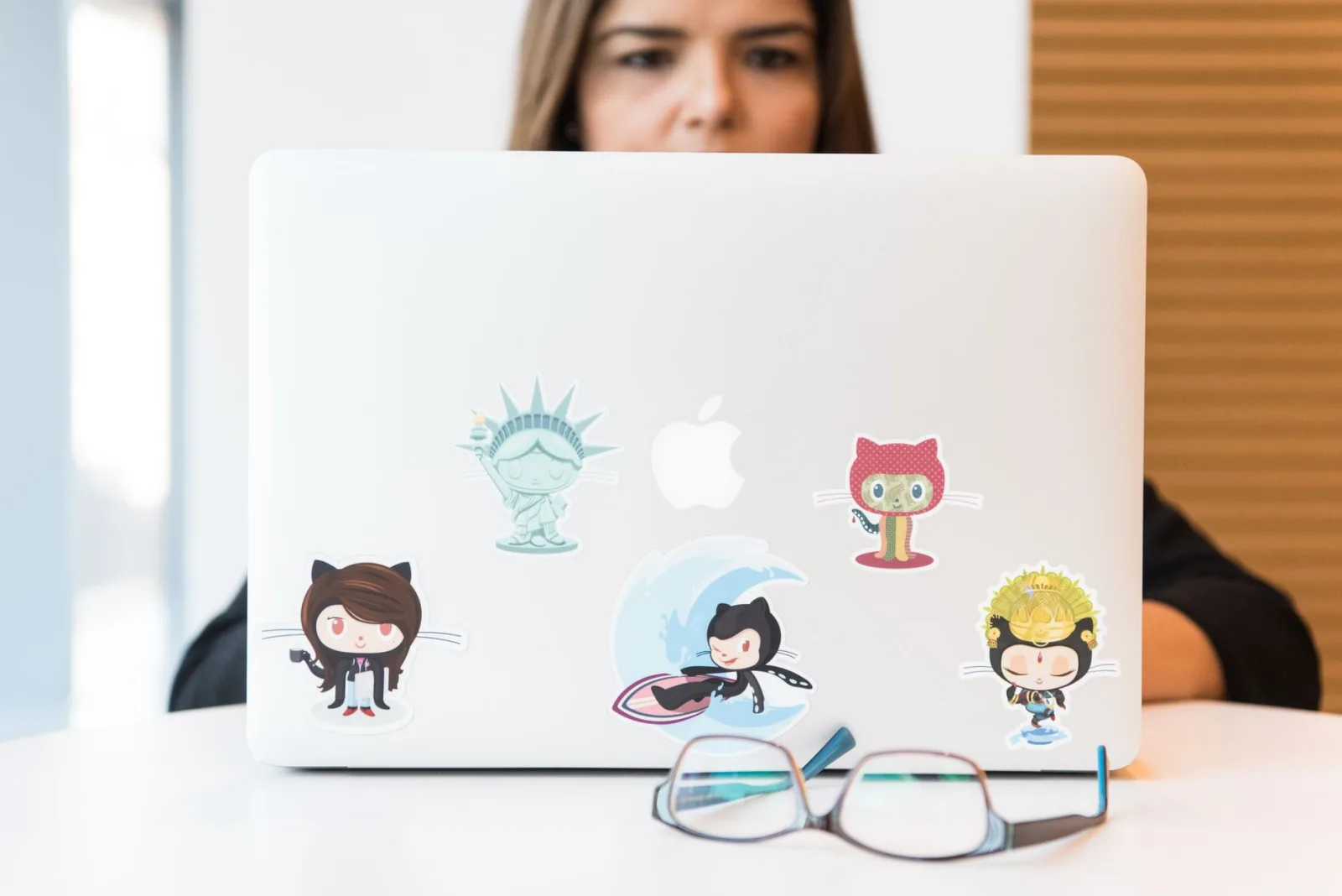 Mujer concentrada en una computadora portátil con pegatinas coloridas con temática de GitHub; gafas en primer plano.