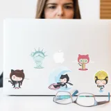 Mujer concentrada en una computadora portátil con pegatinas coloridas con temática de GitHub; gafas en primer plano.