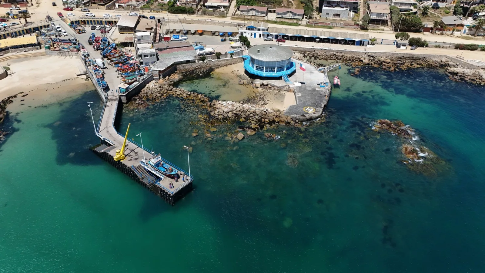 Fotografía aérea del muelle de Algarrobo con barcos pesqueros y edificios costeros, que muestra la vibrante escena costera de Chile.