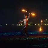 Fascinante actuación de danza con fuego en una playa bajo los cielos nocturnos, con la ciudad de fondo.