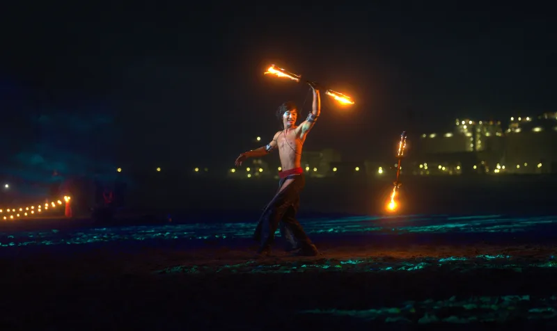 Fascinante actuación de danza con fuego en una playa bajo los cielos nocturnos, con la ciudad de fondo.