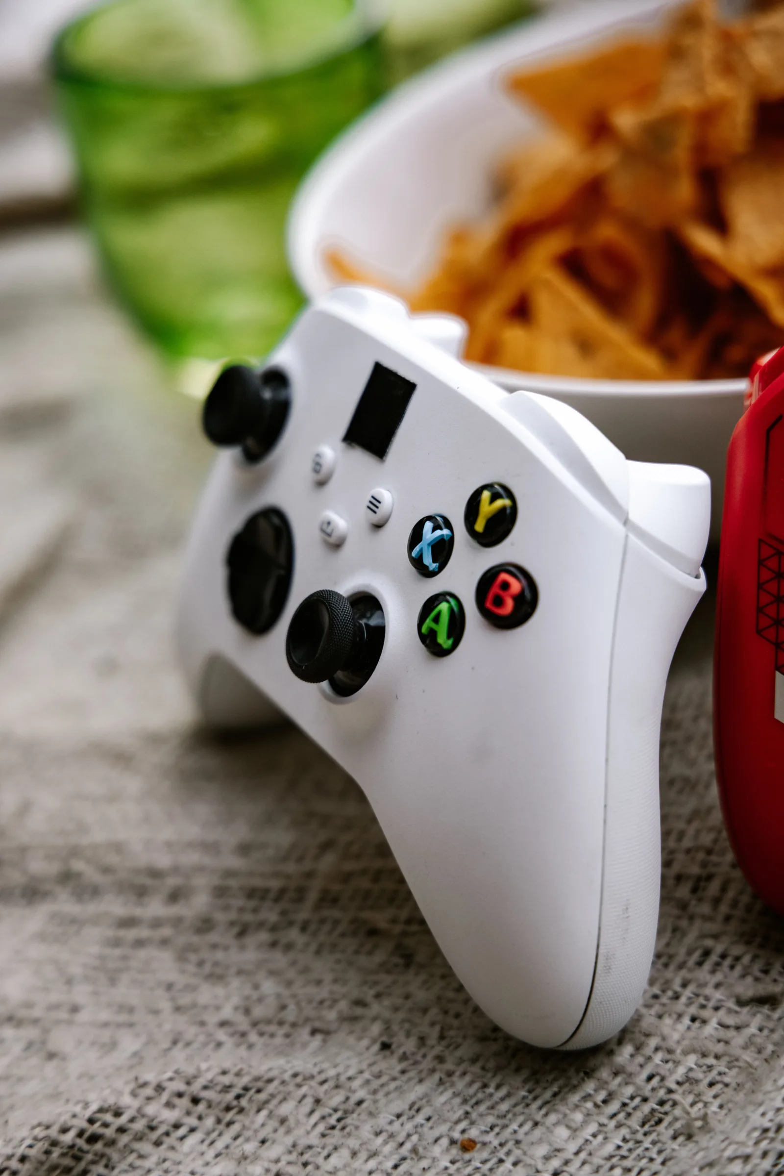 Primer plano de un mando inalámbrico de Xbox junto a un bol de nachos sobre una superficie texturizada.