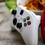 Primer plano de un mando inalámbrico de Xbox junto a un bol de nachos sobre una superficie texturizada.
