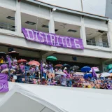 A lively crowd in a stadium stands under umbrellas during a community event on a bright day.