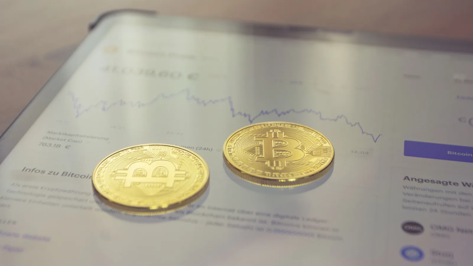 Primer plano de dos monedas doradas de Bitcoin sobre la pantalla de una tableta que muestra datos financieros.